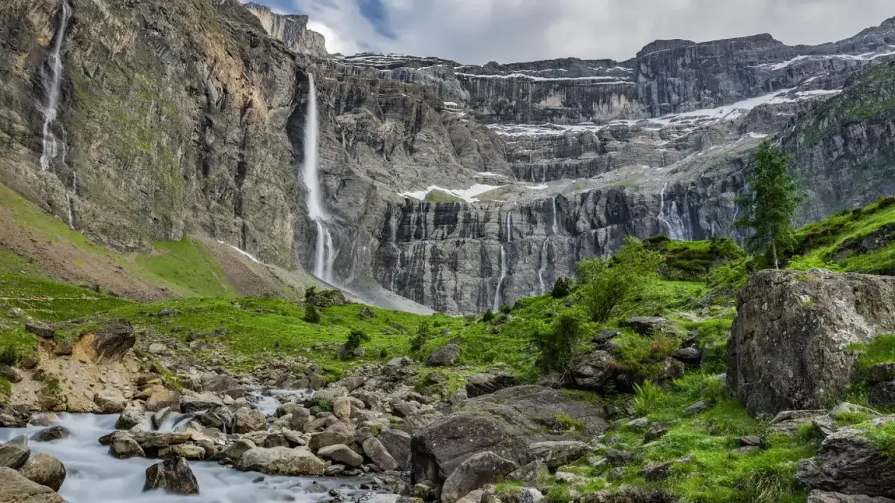 Cascada majestuosa cerca del pueblo de Gavarnie, con rocas y vegetaci&oacute;n verde en primer plano.