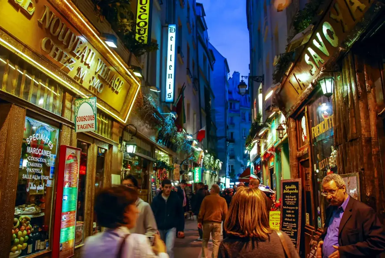 Callej&oacute;n iluminado por la noche, con letreros de restaurantes y gente paseando. Parece un vibrante barrio latino lleno de vida y aromas.