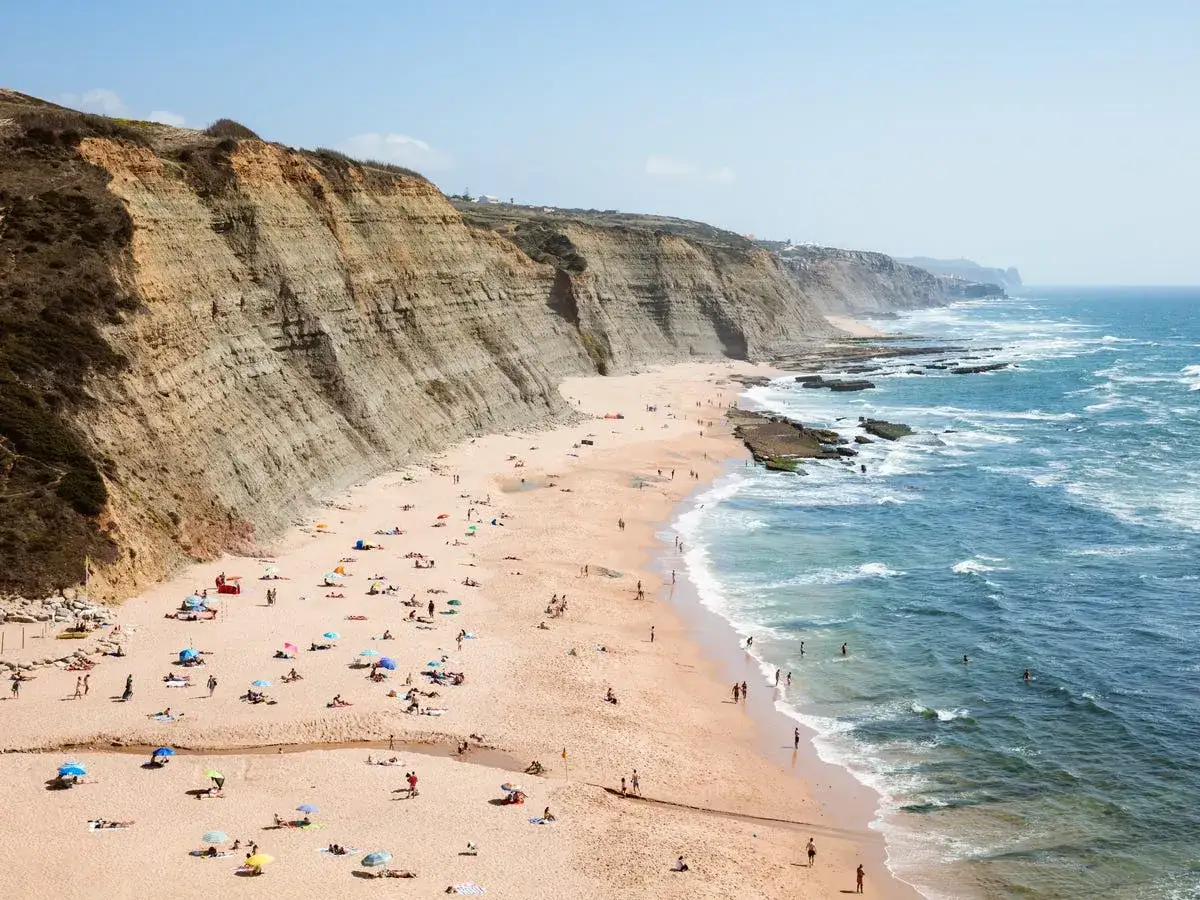 Amplia vista de las **playas de Portugal** con acantilados, arena dorada, gente disfrutando del sol y el mar azul con olas.