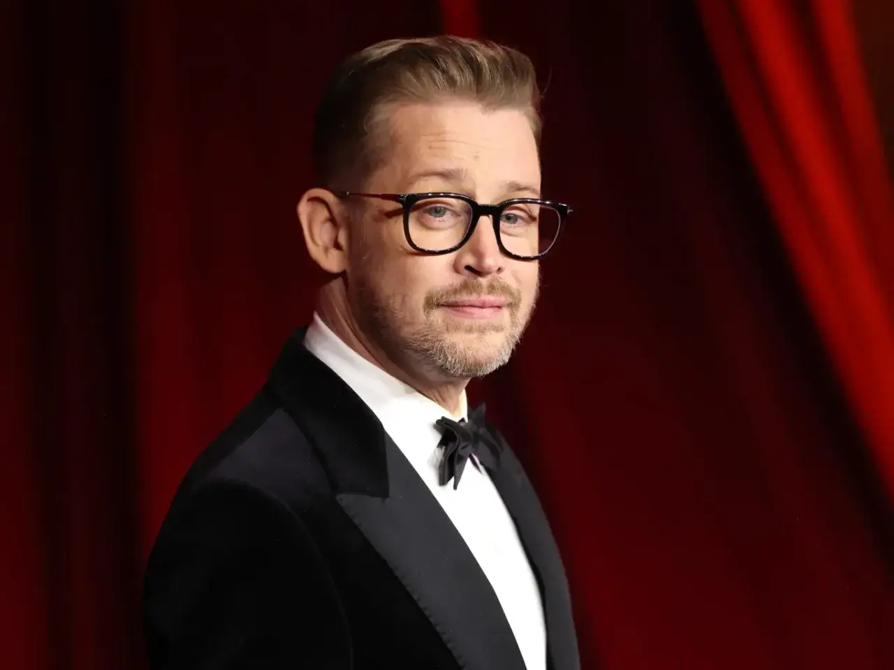Macaulay Culkin w garniturze i okularach, przypomina postać z filmów, w jakich grał.