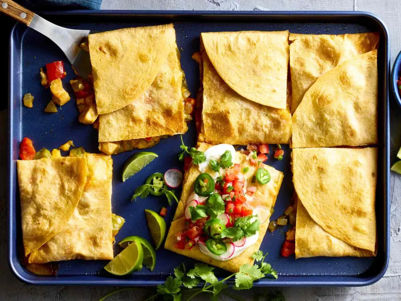 Goldbraune Quesadillas, gef&uuml;llt mit K&auml;se und Gem&uuml;se, werden auf einem blauen Blech serviert. Eine ist mit Pico de Gallo, Sauerrahm und Jalape&ntilde;os garniert.