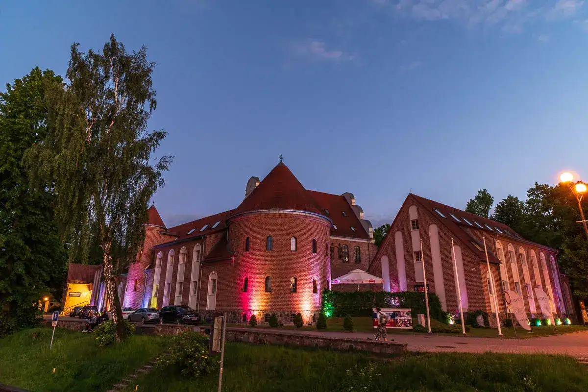 Wieczorem oświetlony zamek w Giżycku, z ceglanymi murami i czerwonymi dachami, prezentuje się malowniczo na tle granatowego nieba.