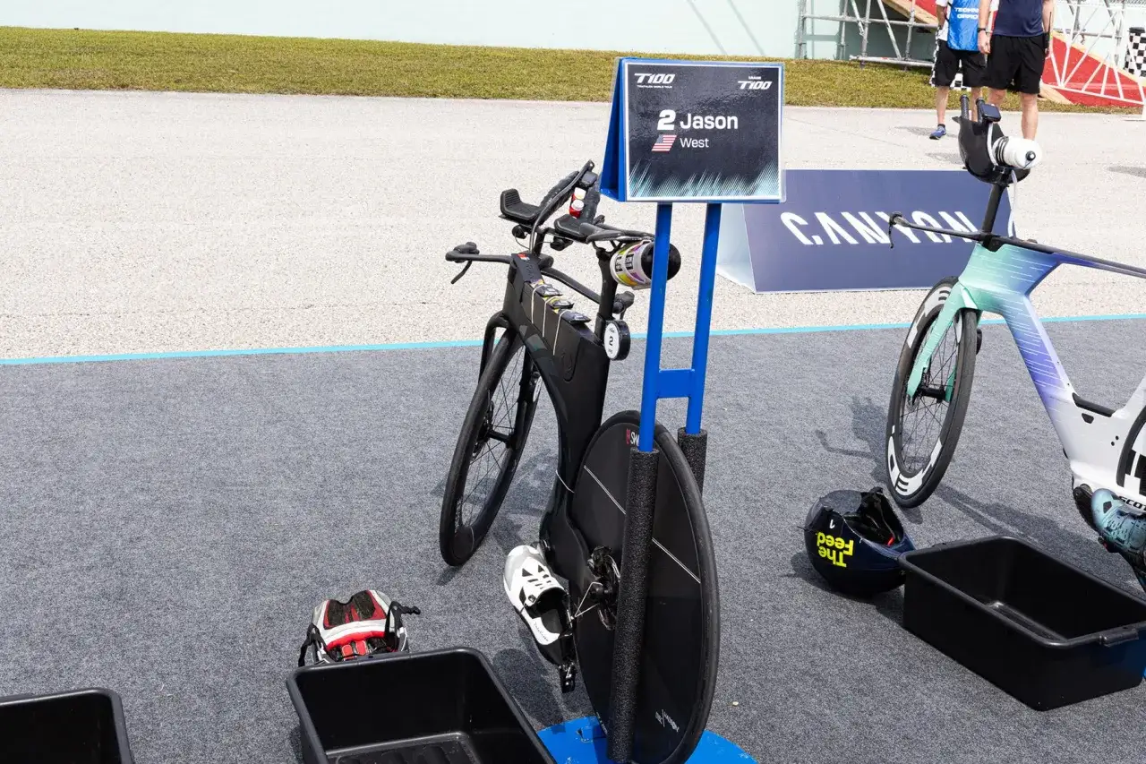 A triathlong transition area with bikes, helmets, and bins. Jason West's bike is prominently displayed with his name tag.