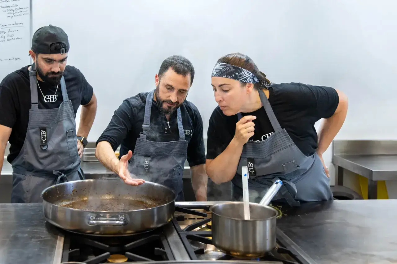 Tres chefs observan atentamente una sartén humeante, evaluando el tiempo de cocción.
