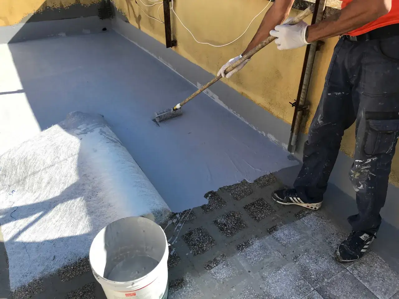 Operaio stende guaina per impermeabilizzazione terrazzo con rullo. Rotolo di tessuto non tessuto e secchio di materiale impermeabilizzante a terra.