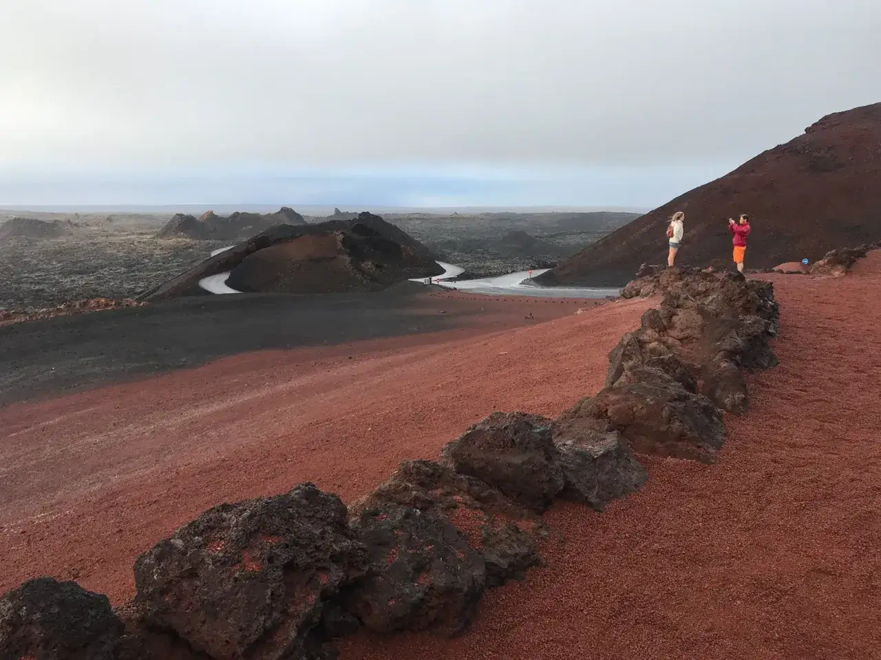 Zwei Personen fotografieren die vulkanische Landschaft im Nationalpark Timanfaya. Rote Erde und dunkle Lava pr&auml;gen das Bild.