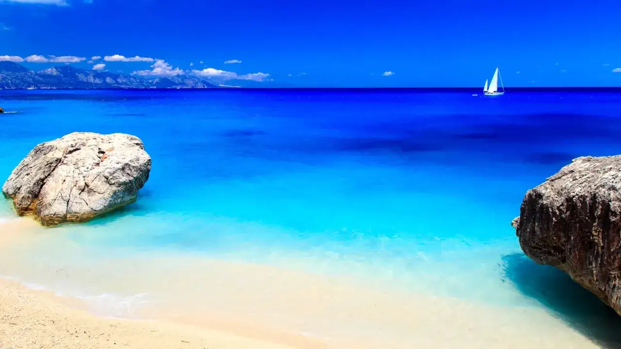 Spiaggia con rocce e mare cristallino, un veliero solca l'orizzonte del mare nord Italia.