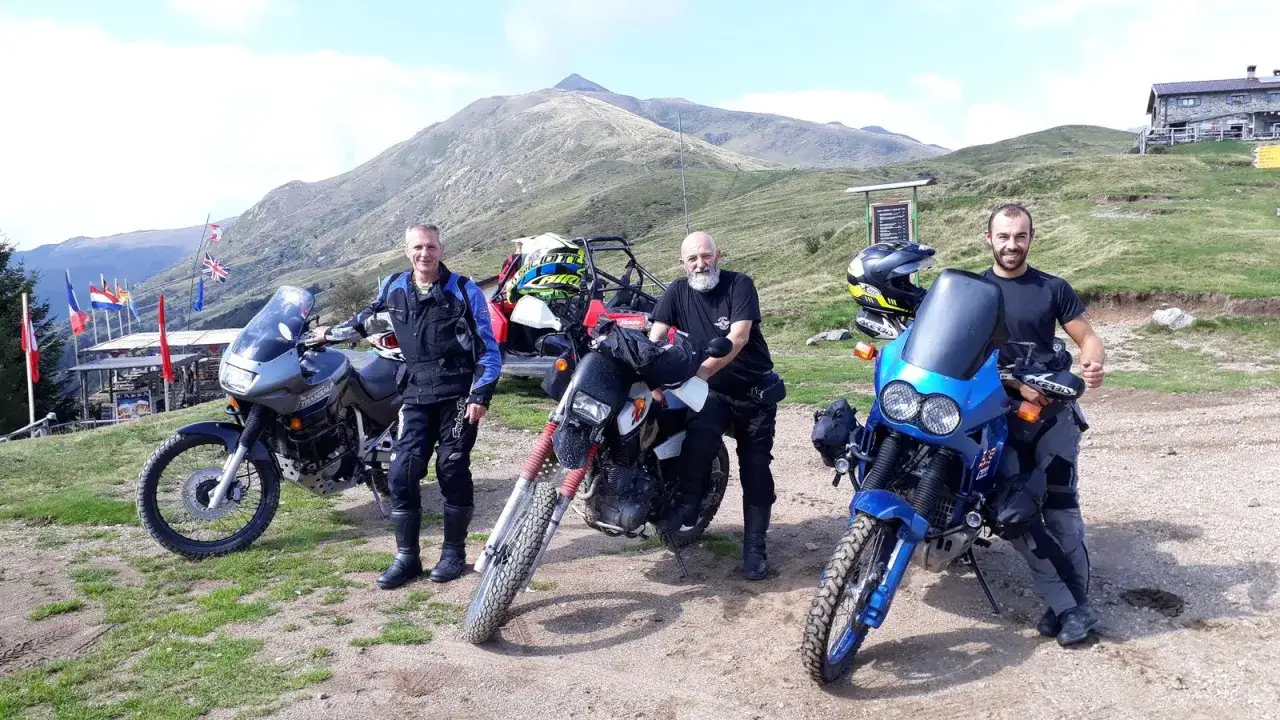 Tre motociclisti con moto da enduro posano al passo San Lucio, con montagne sullo sfondo e un rifugio.