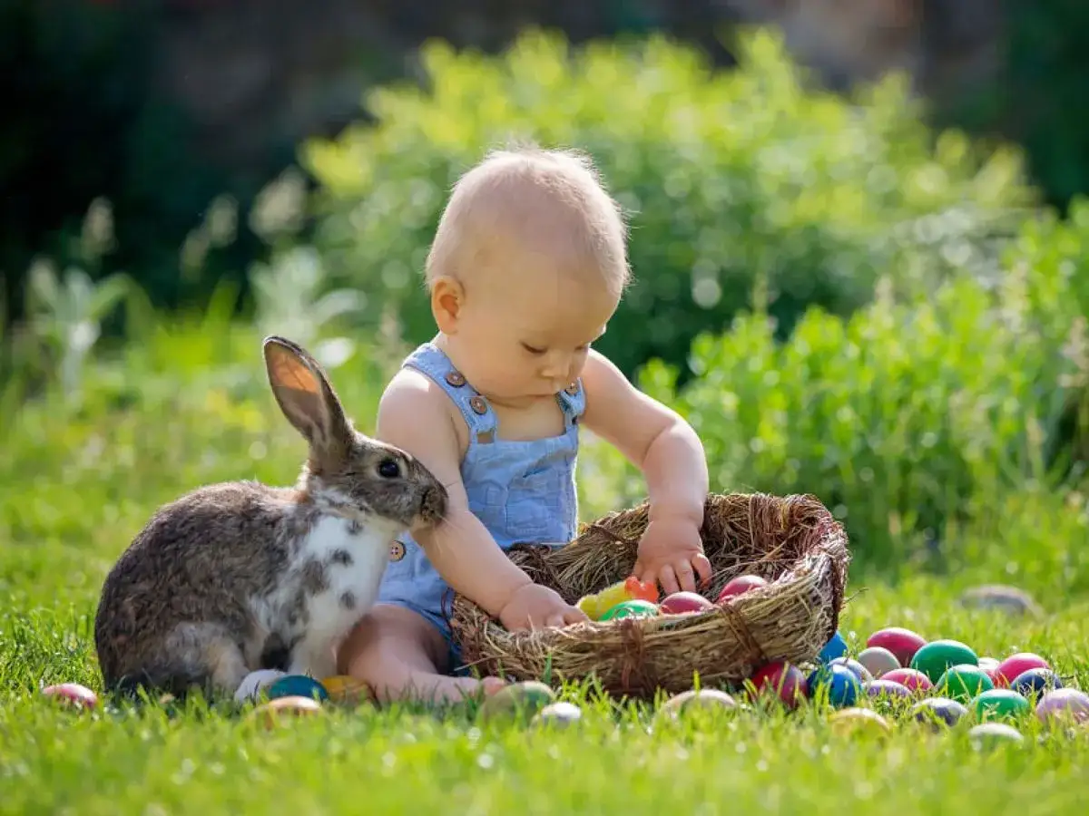 Un bébé et un lapin explorent un panier d'œufs colorés, prêts pour la chasse aux œufs de Pâques dans l'herbe verte.