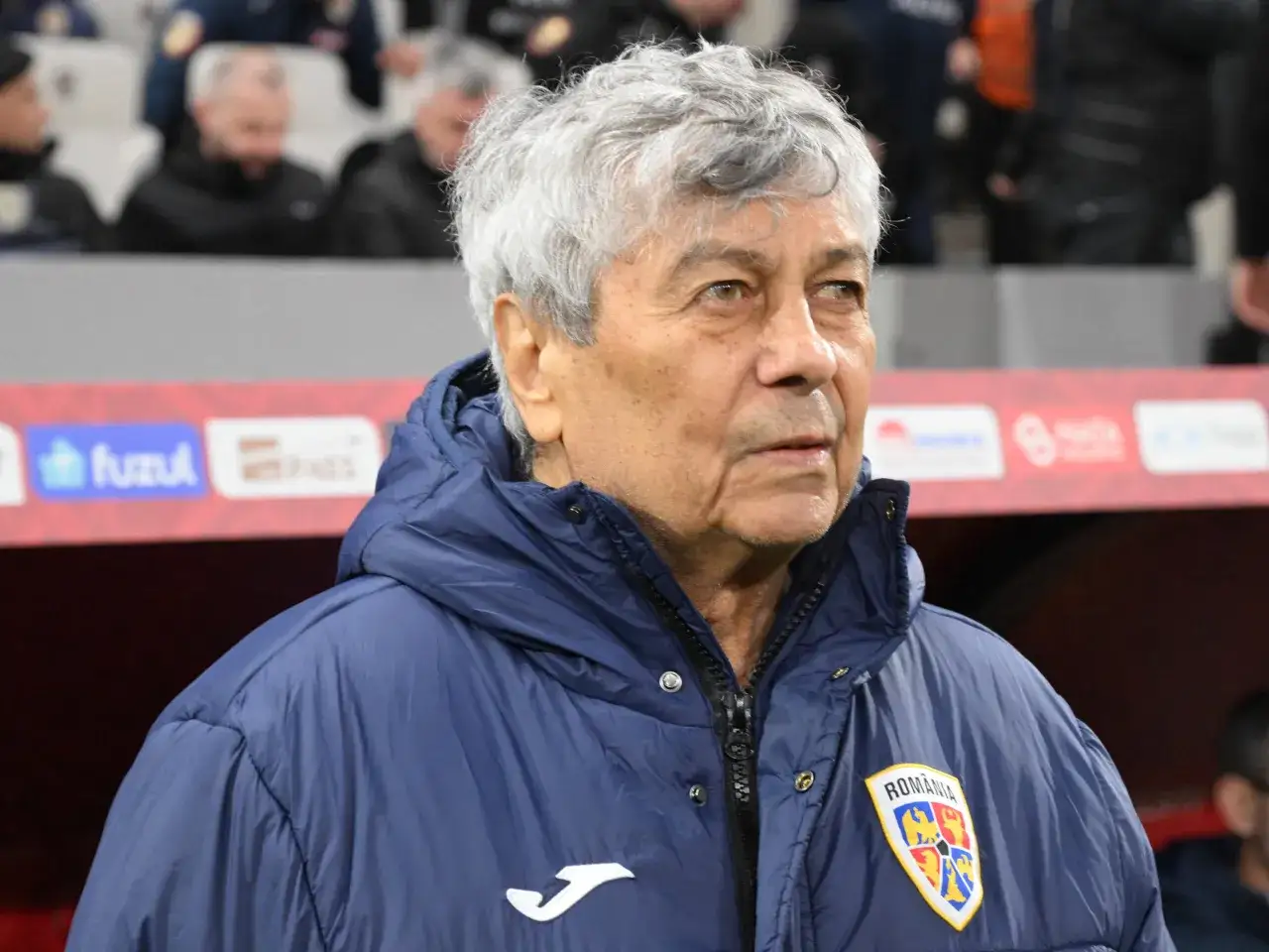 Entrenador con pelo gris, viste chaqueta azul con escudo de Rumanía. Podría ser Carlo Ancelotti en un partido.