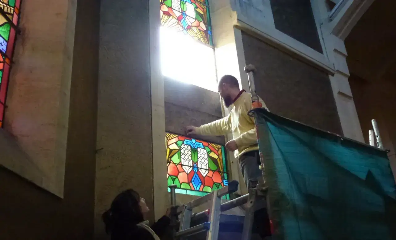 Un homme sur un échafaudage ajuste un vitrail coloré dans une église. Une femme regarde.