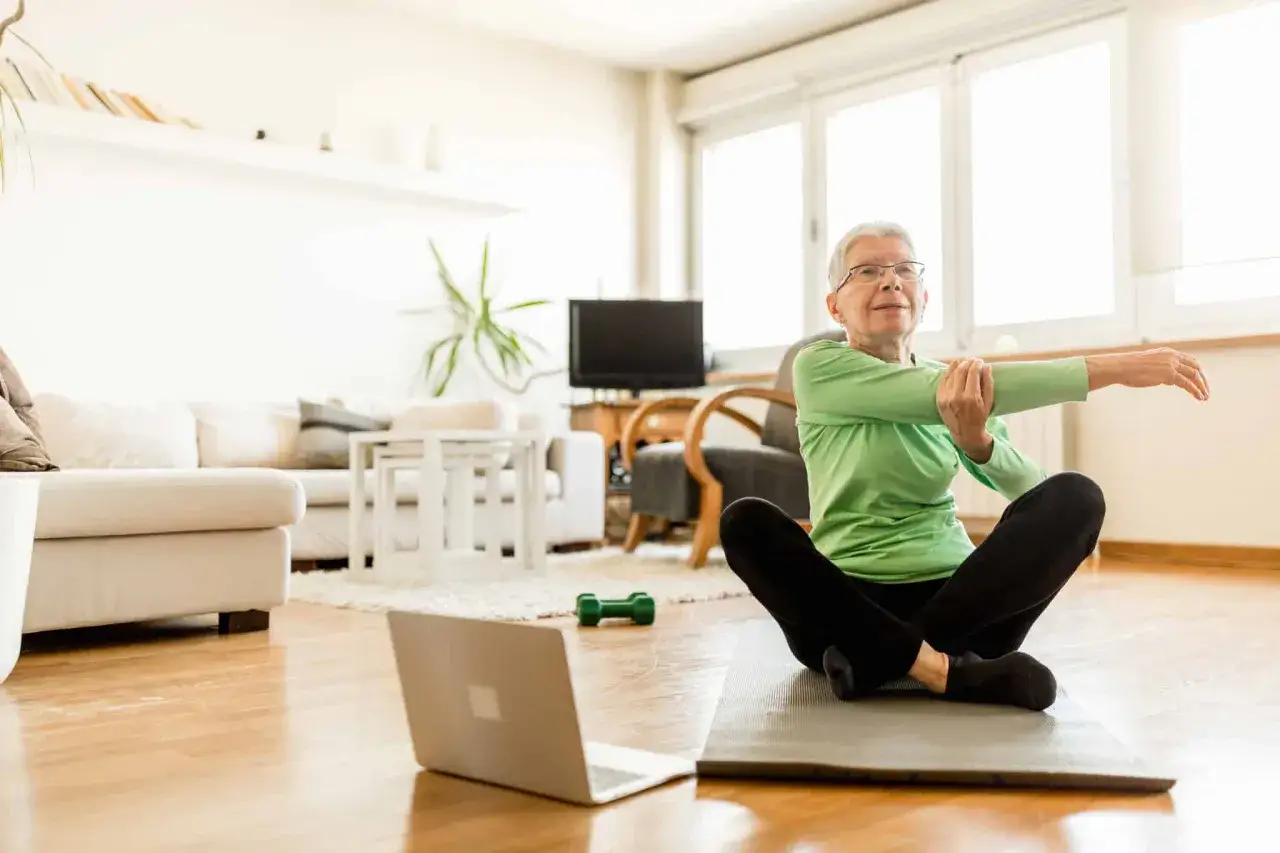 Une femme &acirc;g&eacute;e fait du sport chez soi, assise sur un tapis, suivant une vid&eacute;o sur son ordinateur portable.
