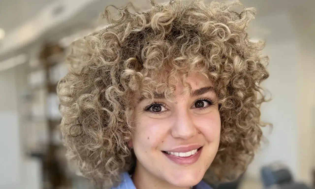 Mujer sonríe con un espectacular pelo rizado con flequillo rubio.