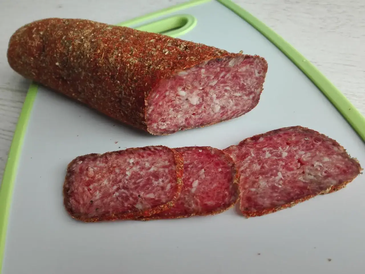 Kawałek wiejskiej kiełbasy, z czego jest salami, pokryty przyprawami, leży na desce. Obok trzy plastry.