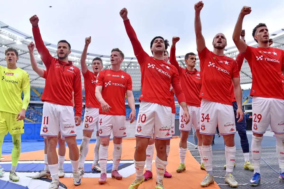 Piłkarze Wisły Kraków w czerwonych strojach świętują, podnosząc ręce. Wszyscy czekają na nowe transfery, by wzmocnić drużynę.