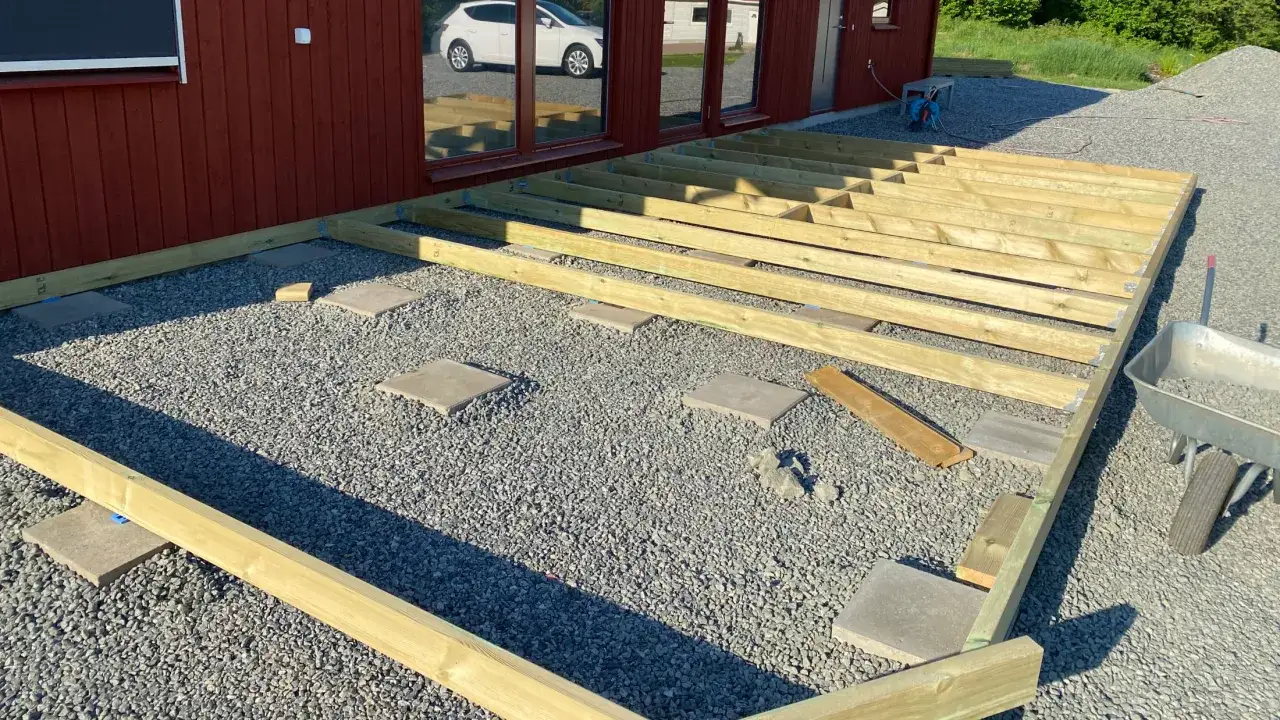 Grundarbete för att bygga altan på plattor. Träreglar ligger redo att monteras på grus och stenplattor.