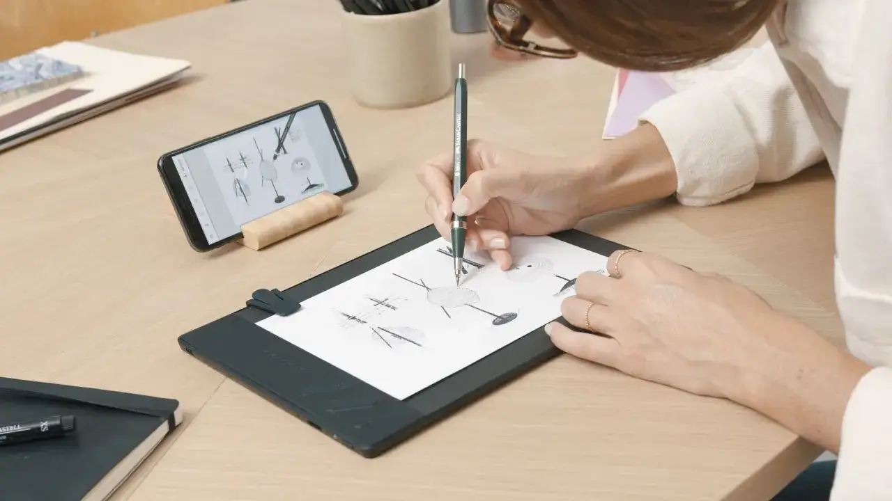 Kobieta szkicuje na papierze przy użyciu tabletu graficznego bez podłączania do komputera. Obok leży smartfon z inspiracjami.