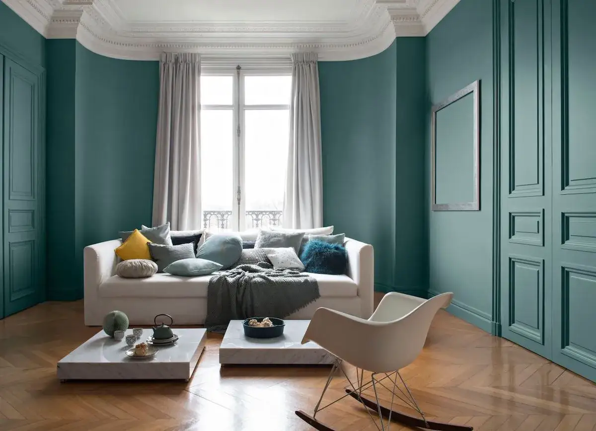 Salon aux murs bleu ou vert canard, canapé blanc garni de coussins, tables basses en marbre, fauteuil à bascule et parquet.