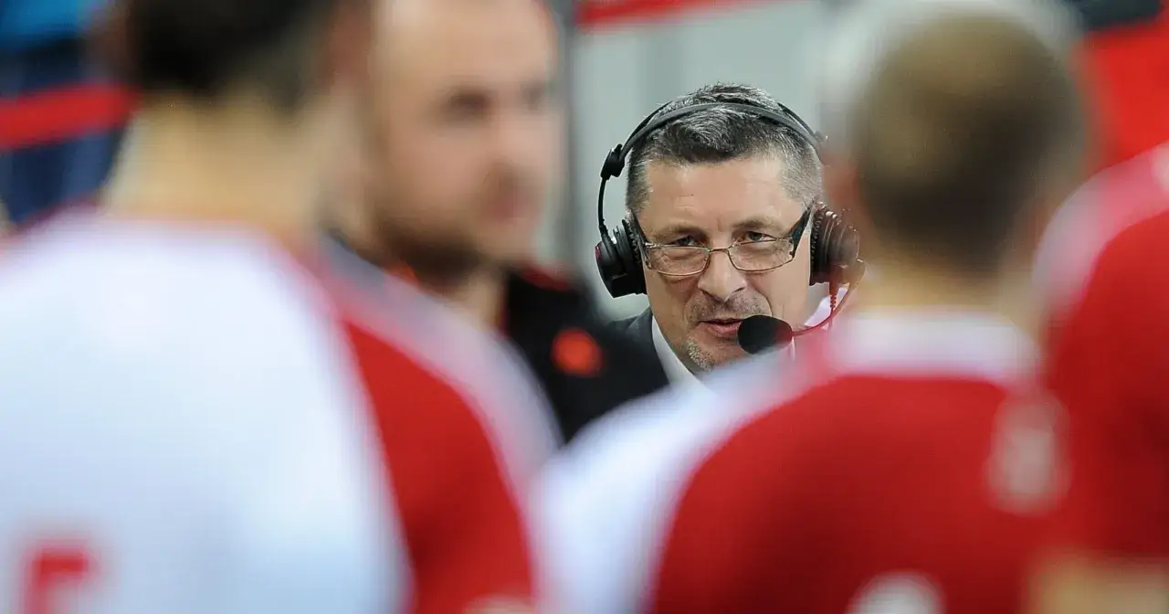 Ireneusz Mazur w słuchawkach z mikrofonem, otoczony przez zawodnik&oacute;w w czerwonych strojach.