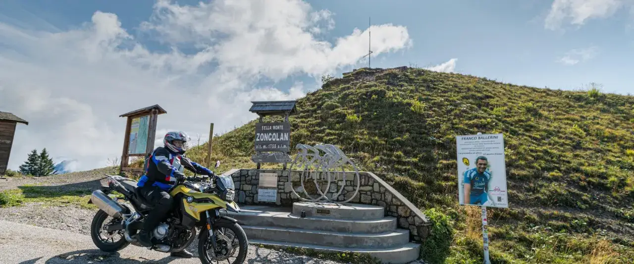 Motorradfahrer am Monte Zoncolan, mit Schildern und einer Radfahrer-Skulptur.