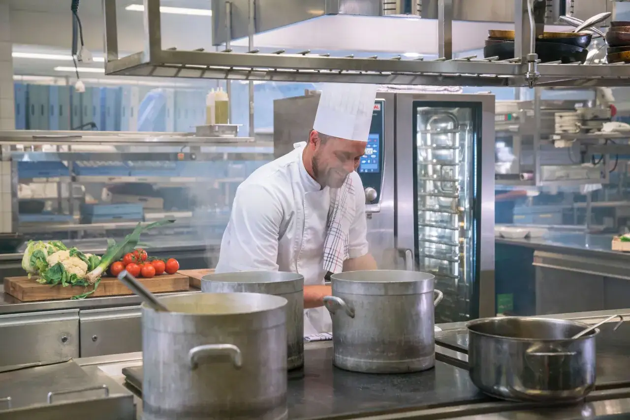 En leende su chef i vitt kökskläder arbetar vid spisen med stora kastruller. Grönsaker ligger på en skärbräda.