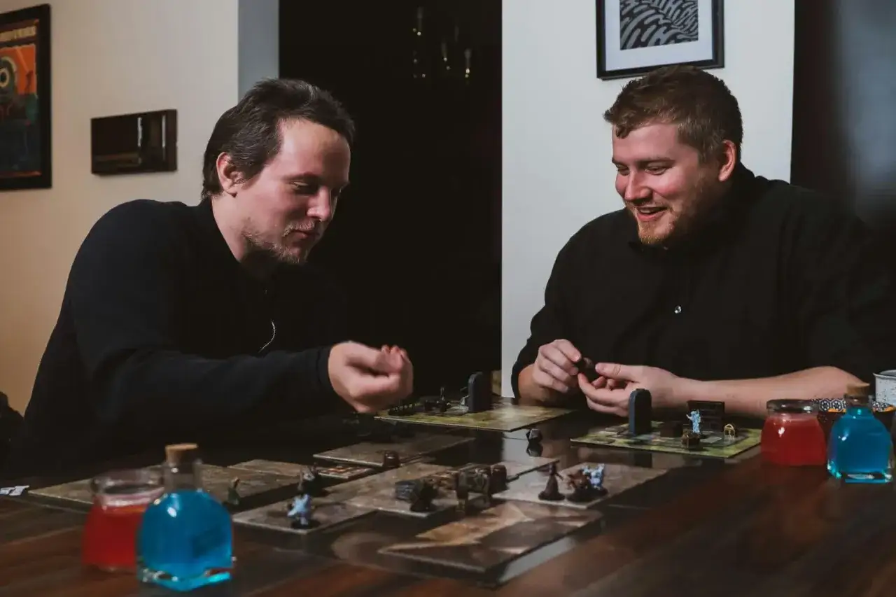 Två män spelar ett brädspel med figurer och tärningar. De skrattar och verkar ha roliga utmaningar på jobbet, trots att det ser ut som en fritidsaktivitet.
