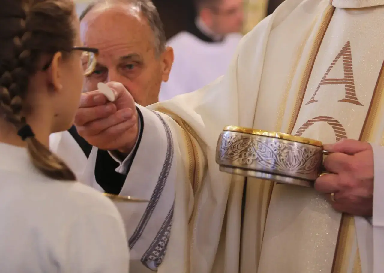 Kapłan podaje dziewczynce białą hostię podczas komunii świętej. W ręku trzyma ozdobny kielich.