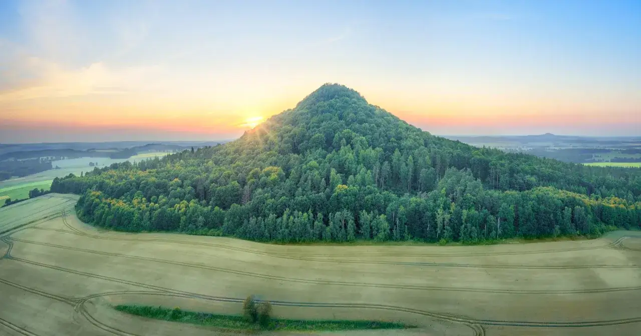 Wzgórze porośnięte lasem, przypominające kształtem wygasły wulkan, w krainie wygasłych wulkanów. Zachodzące słońce oświetla zbocze.