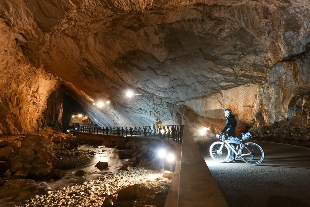Ciclista esplora i più bei percorsi in bicicletta in Italia, attraversando una grotta illuminata con un fiume sotterraneo.