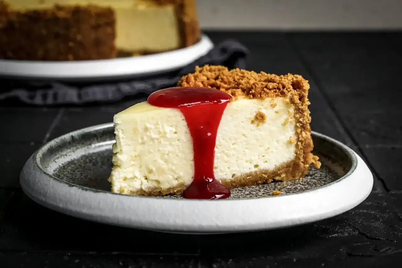 Ein Stück cremiger american cheesecake mit Keksboden, übergossen mit roter Fruchtsauce, auf einem Teller.