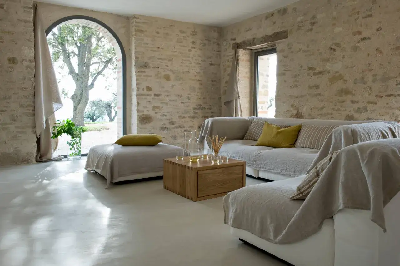 Elige el color del suelo de una casa con este salón rústico. Suelos de microcemento claro y paredes de piedra.