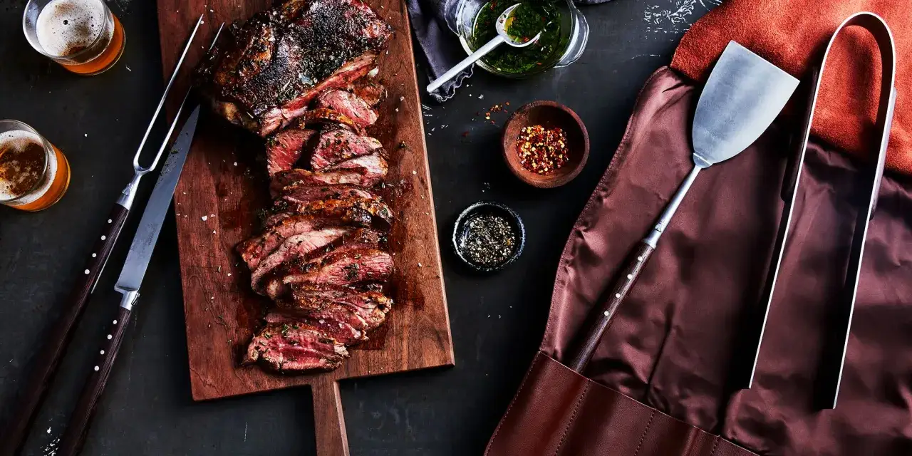 Saftiges, aufgeschnittenes Fleisch zum Grillen liegt auf einem Holzbrett, daneben Bier, Gew&uuml;rze und Grillbesteck.