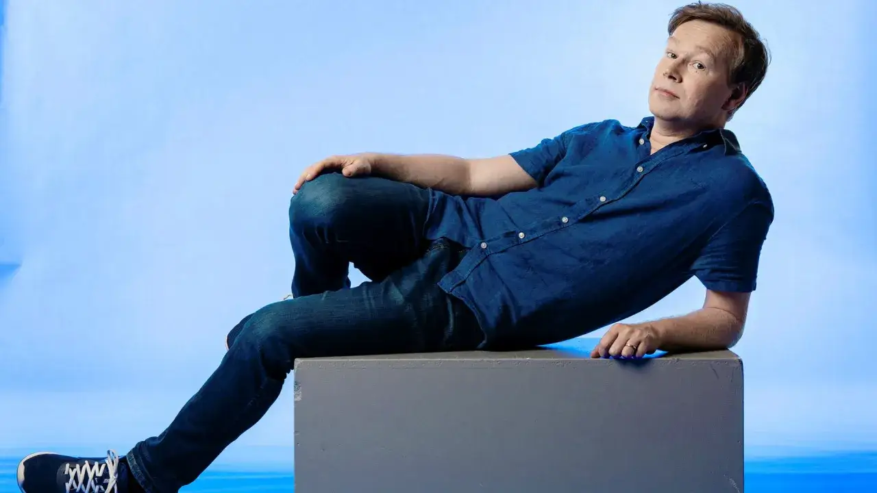 Johan Glans lutar sig avslappnat mot en gr&aring; kub, kl&auml;dd i jeans och en bl&aring; skjorta, med en blick som antyder ett sk&auml;mt.