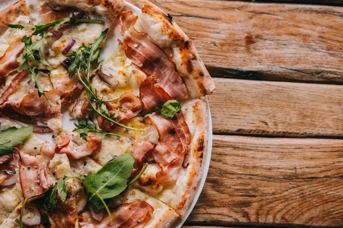 Pyszna pizza carbonara z chrupiącym boczkiem, rukolą i cebulą na drewnianym stole.