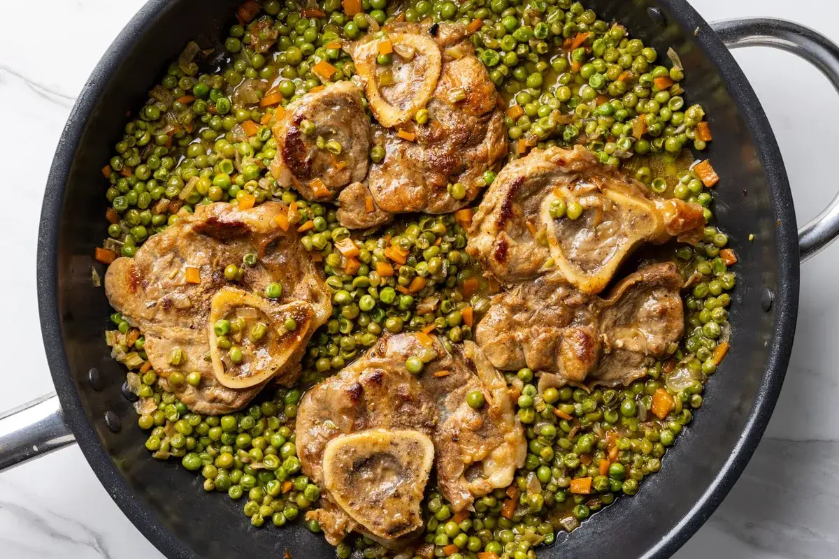 Quattro fette di ossobuco con piselli e carote in padella, un piatto succulento pronto per essere gustato.