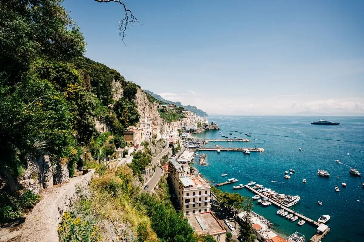 A scenic view of the Amalfi Coast, Italy, with buildings clinging to cliffs and boats in the turquoise sea. This is the legendary walk of the gods.