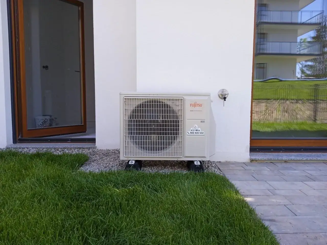 Jednostka zewnętrzna klimatyzatora Fujitsu stoi na trawniku obok białego budynku.