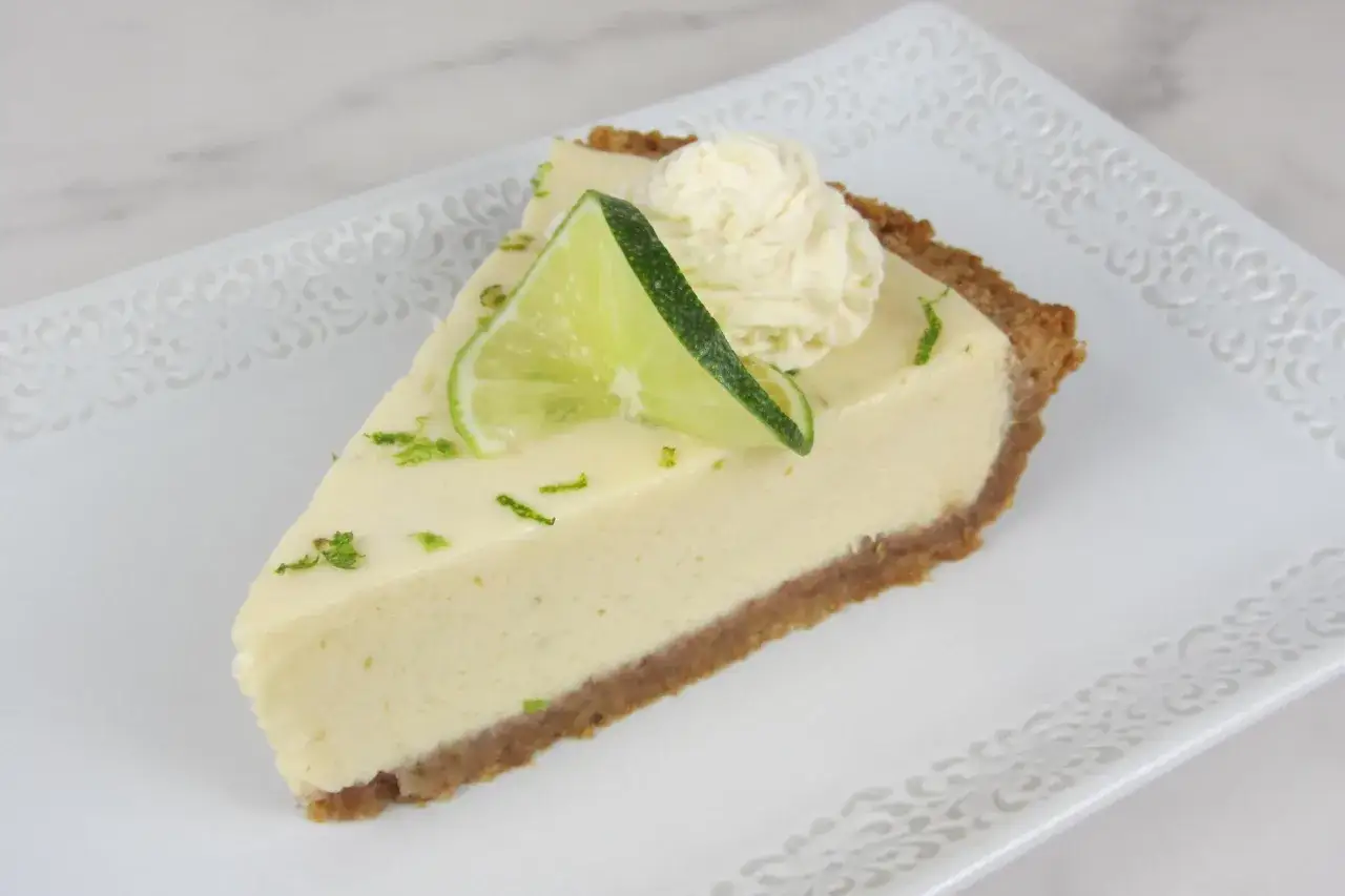 Ein St&uuml;ck cremiger Key Lime Pie mit Limettenscheibe und Sahnetupfer auf einem verzierten Teller.