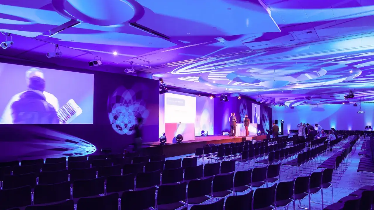 Salle de conférence illuminée en violet, avec des rangées de sièges vides. Le nombre de places est impressionnant pour cet événement de maroquinerie.