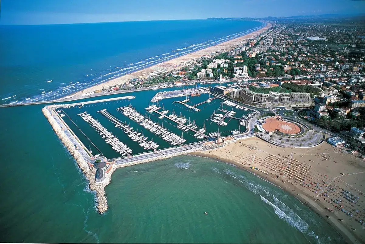Vista aerea del porto turistico di San Giuliano Mare a Rimini, con barche ormeggiate e la spiaggia affollata.