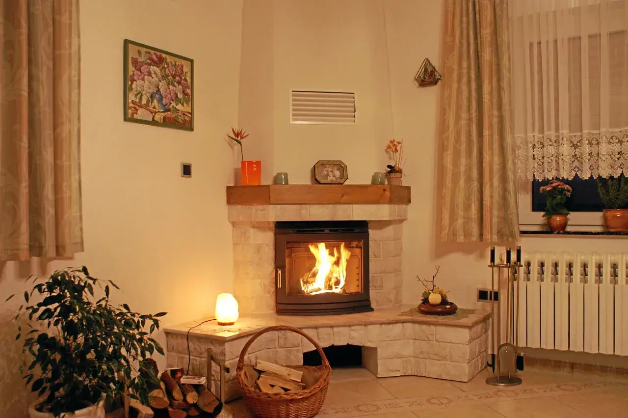 Camino in pietra con fuoco acceso, dimostrando che la calce resiste alle alte temperature. Cesto di legna, pianta e quadro decorano l'ambiente.