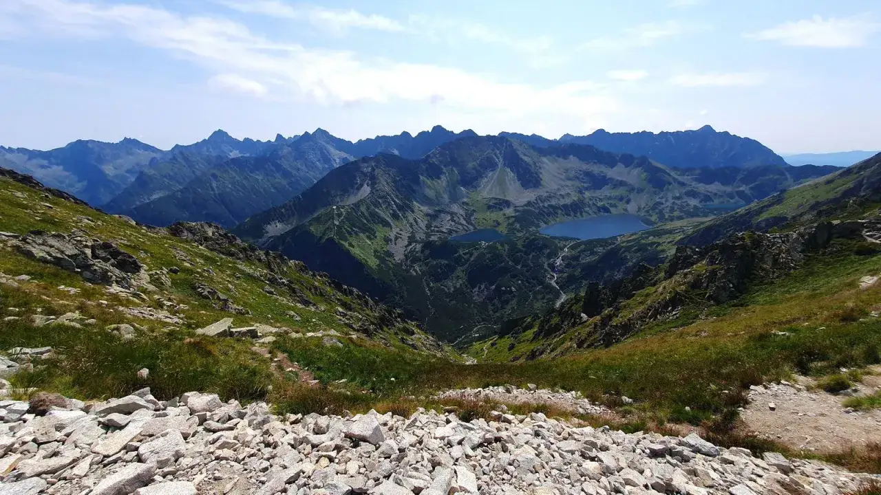 Widok na Tatry z kamienistego zbocza. W dole jeziora i kręte szlaki na krzyżne, otoczone zielenią i skalistymi szczytami.