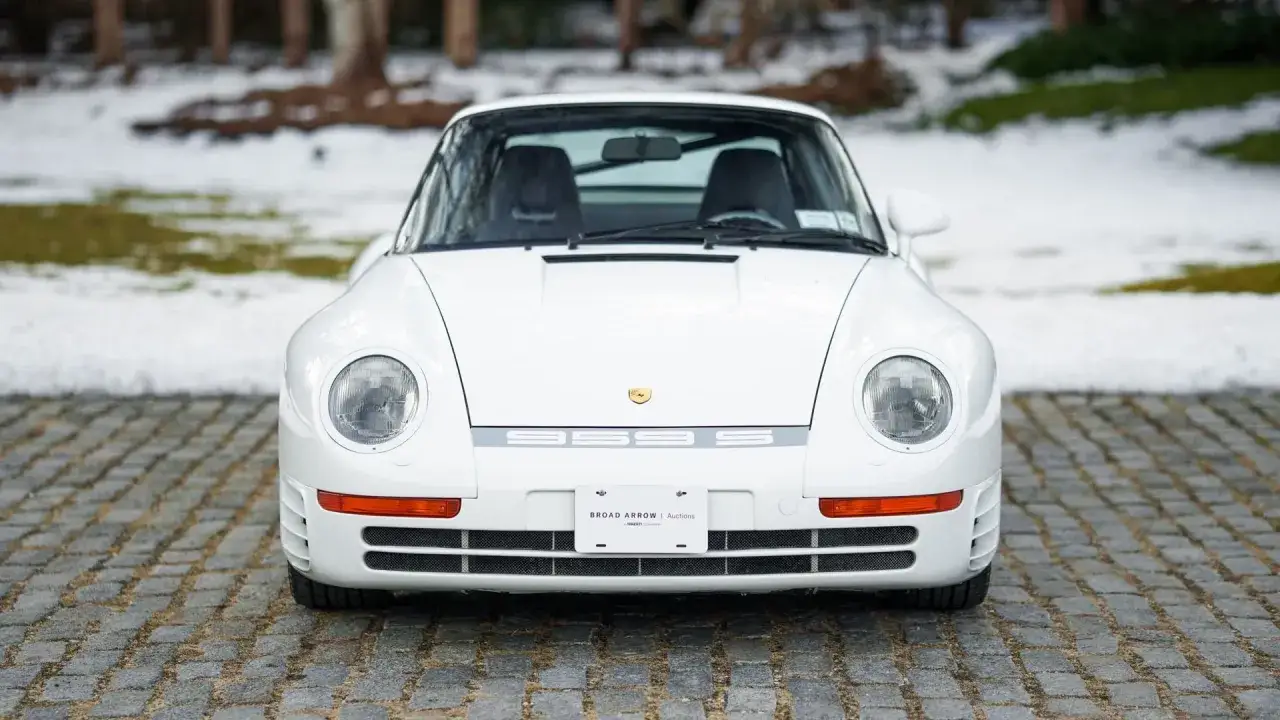 Biały Porsche 959 z lat 80. na brukowanym podjeździe, z tyłu śnieg i drzewa. Ikona motoryzacji.
