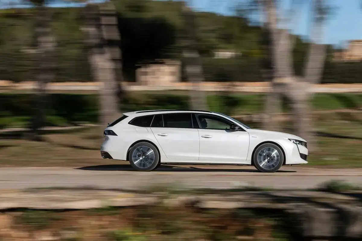 Vit Peugeot 508 SW i fart under ett test. Bilen glider fram längs en väg med suddig bakgrund av träd och stenar.