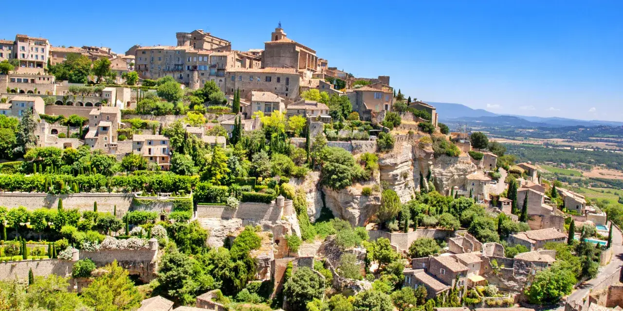 Village perch&eacute; de Gordes, avec ses maisons en pierre et ses jardins luxuriants, &eacute;voquant l'ambiance d'un march&eacute; proven&ccedil;al anim&eacute;.
