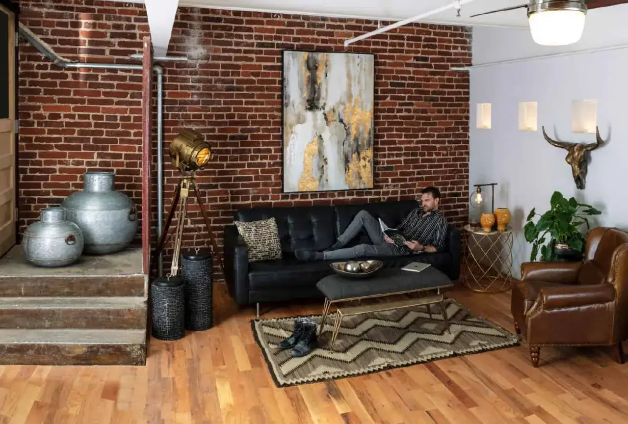 Gem&uuml;tliches **vintage industrial style wohnzimmer** mit Mann auf schwarzem Sofa, Ziegelwand, Metallakzenten und Holzboden.