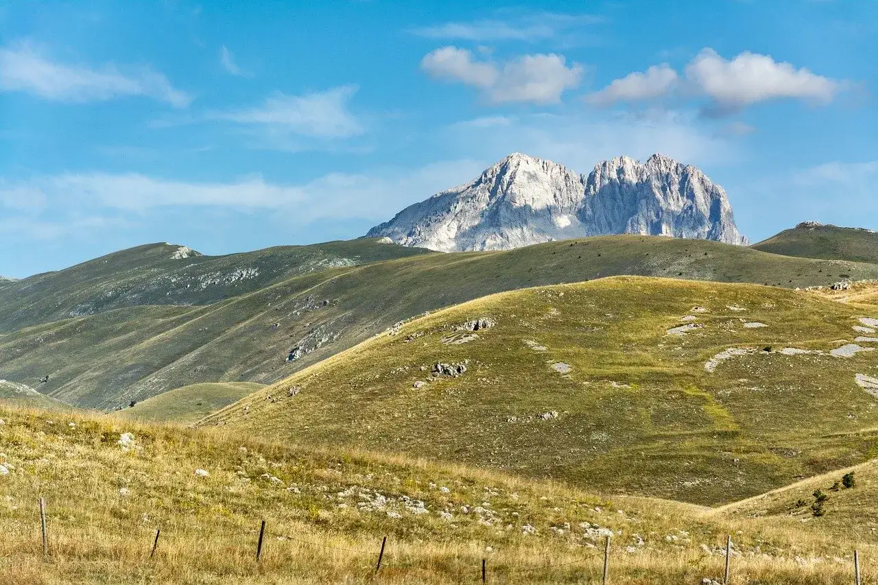 Vaste colline verdi e dorate sotto un cielo azzurro con nuvole bianche, con montagne imponenti sullo sfondo. Un paesaggio che evoca il **cammino dei briganti in Abruzzo**.