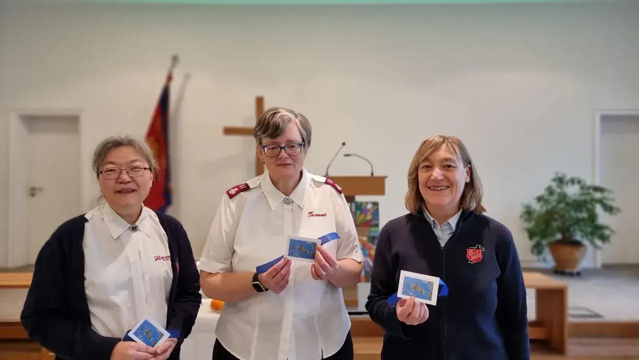 Drei Frauen der Heilsarmee halten kleine Karten mit blauen Bändern. Sie tragen Uniformen der Heilsarmee.