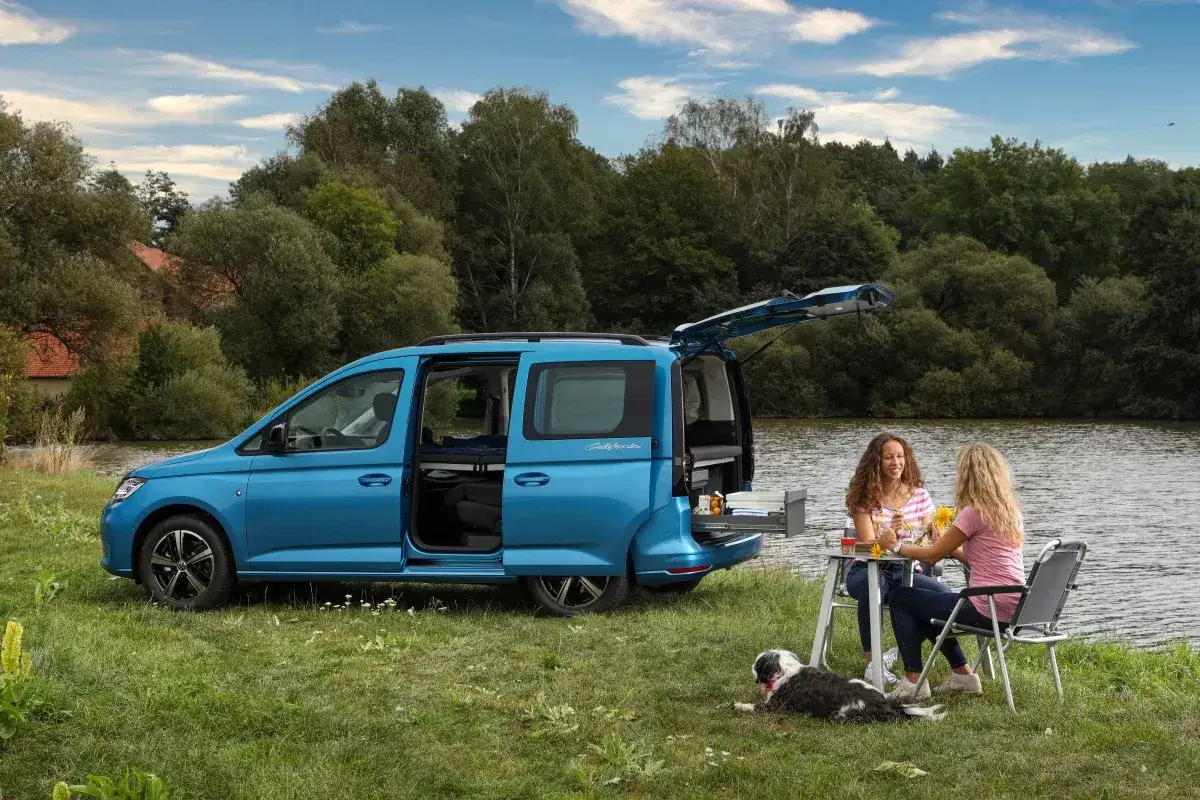 Niebieski caddy kamper z otwartymi drzwiami i bagażnikiem, przy którym siedzą dwie kobiety i pies.