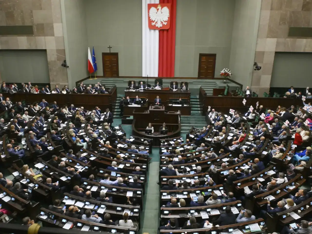 Wielka sala obrad, gdzie odbywa się **zgromadzenie narodowe**. Posłowie siedzą w ławach, a na mównicy przemawia jeden z nich.